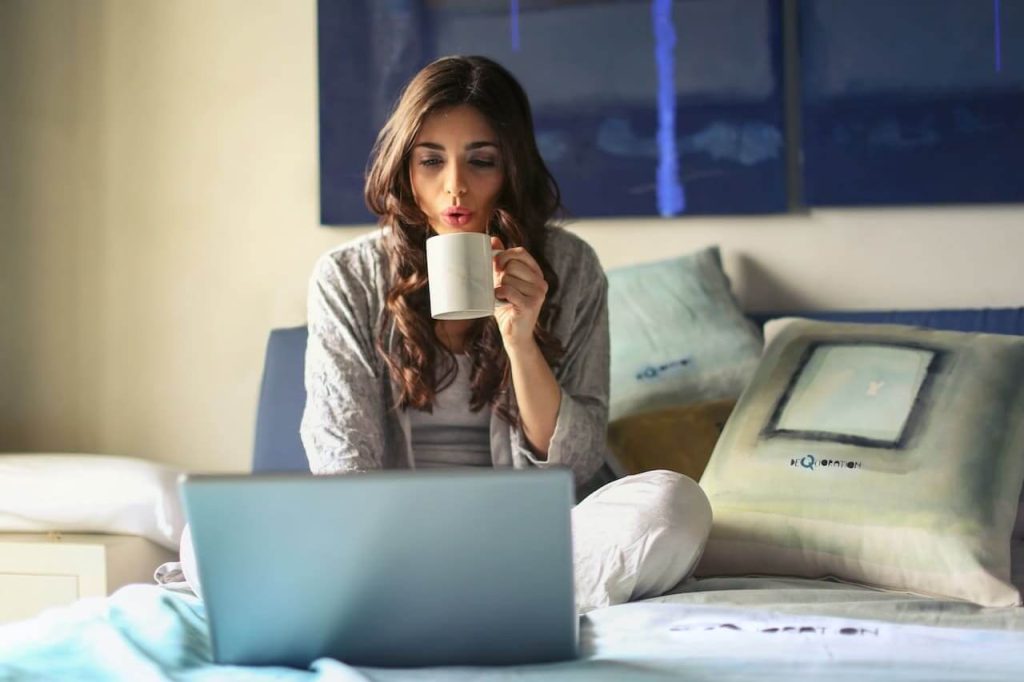 Trabalho home office: mulher sentada na cama olhando para o notebook e tomando uma xícara de café.