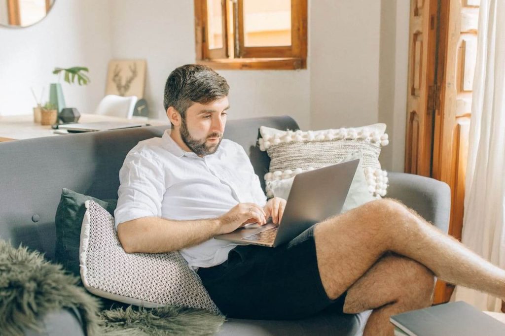 Vagas home office: homem de bermuda sentado no sofá com notebook no colo.